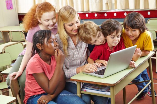 Kinder sitzen mit ihrer Erzieherin am Tisch und schauen auf ein Laptop